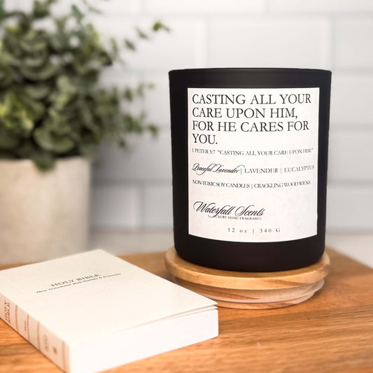 Christian scripture candle in a black glass jar with white label showing 1 Peter 5:7. Hand poured soy wax with a wooden wick for peaceful ambiance.