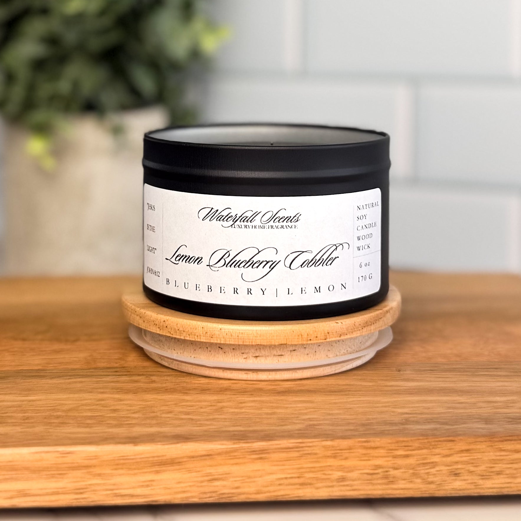 Lemon Blueberry Cobbler wooden wick candle in a black travel tin with a white label. A cozy, non-toxic candle with a blend of warm lemon, sweet blueberries, and sugar crumble.