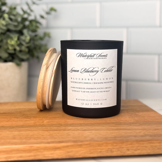 Lemon Blueberry Cobbler wooden wick candle in a black glass jar with a white label. Hand poured, non-toxic, clean burning, with scents of lemon zest, ripe blueberries, and sugar.